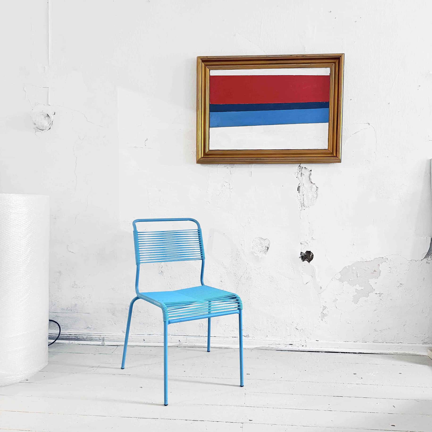 Blaue Metallstuhl vor weißer Wand mit moderner abstrakter Kunst im Goldrahmen, geschaffen von Nikolaus Kriese, im Rahmen seiner Malerei & Auftragsmalerei.