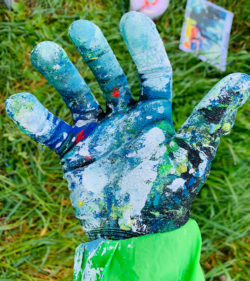 Farbige, bemalte Hand mit Spritzern und Texturen, auf grünem Ärmel, Landleben im Hintergrund, kreative Malerei, Auftragsmalerei, Künstlerarbeit, Outdoor-Kunst.