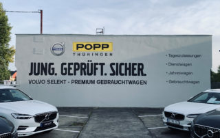 Bilderwand mit Werbung für Volkswagen Gebrauchtwagen in Thüringen, zeigt verschiedene Modelle, Parkplatz und Werbetafel mit Slogans und Serviceangeboten, ideal für Auto- und Fahrzeugfotografie.