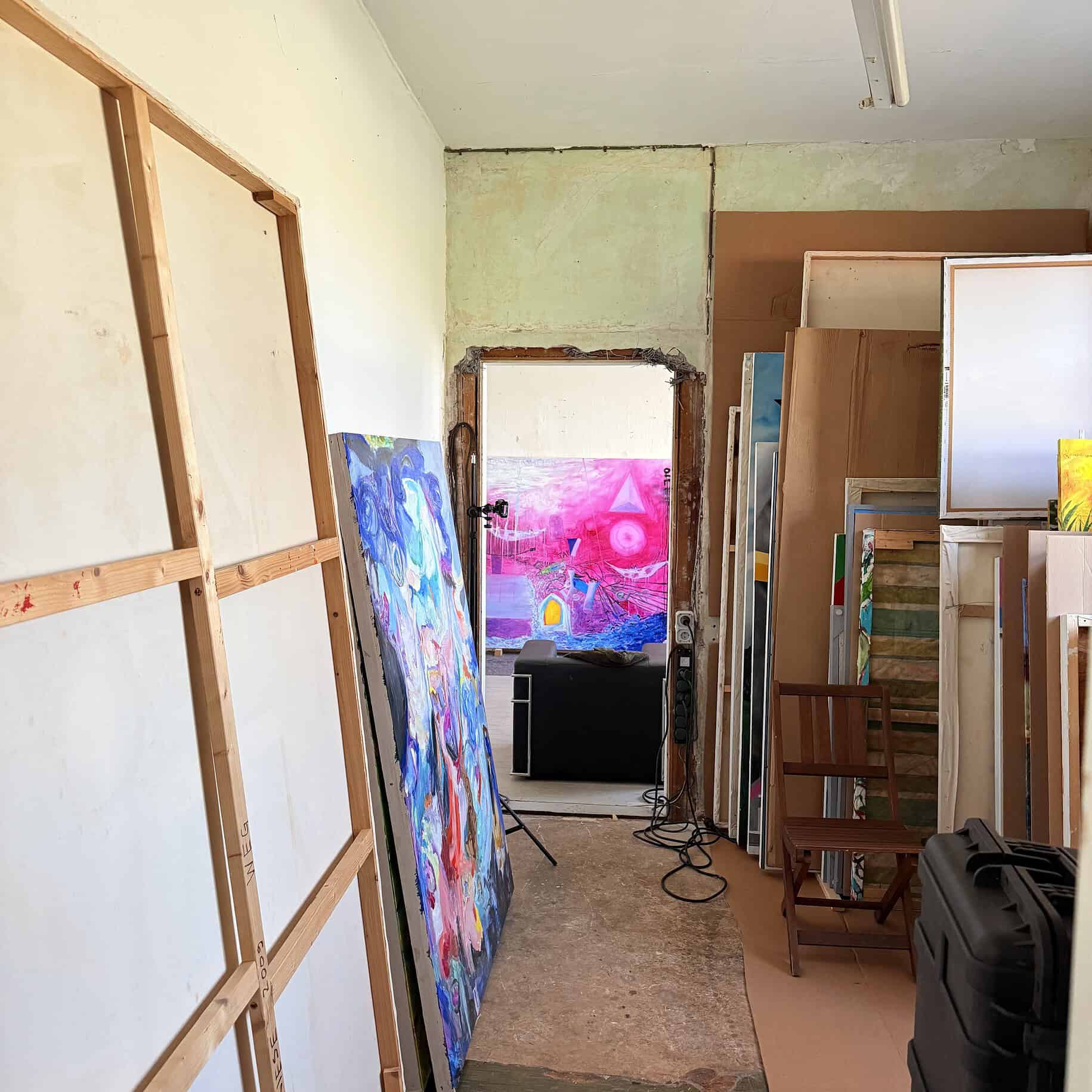 A view inside a painting studio with several abstract paintings hanging on the walls and standing on easels; in the background, a colorful abstract painting.