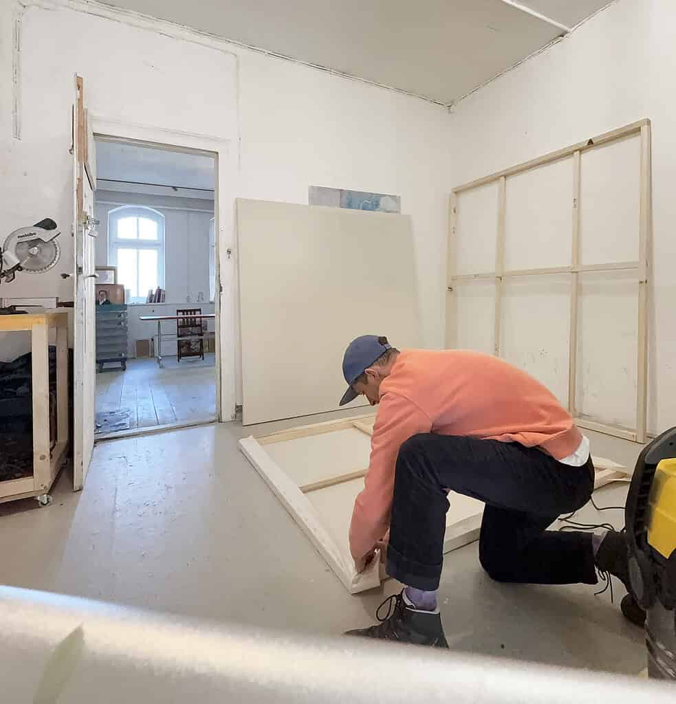 Künstler beim Aufbau eines Rahmens in einem Atelier, Vorbereitung für ein Gemälde, in einem hellen Raum mit weißen Wänden und großen Fenstern.