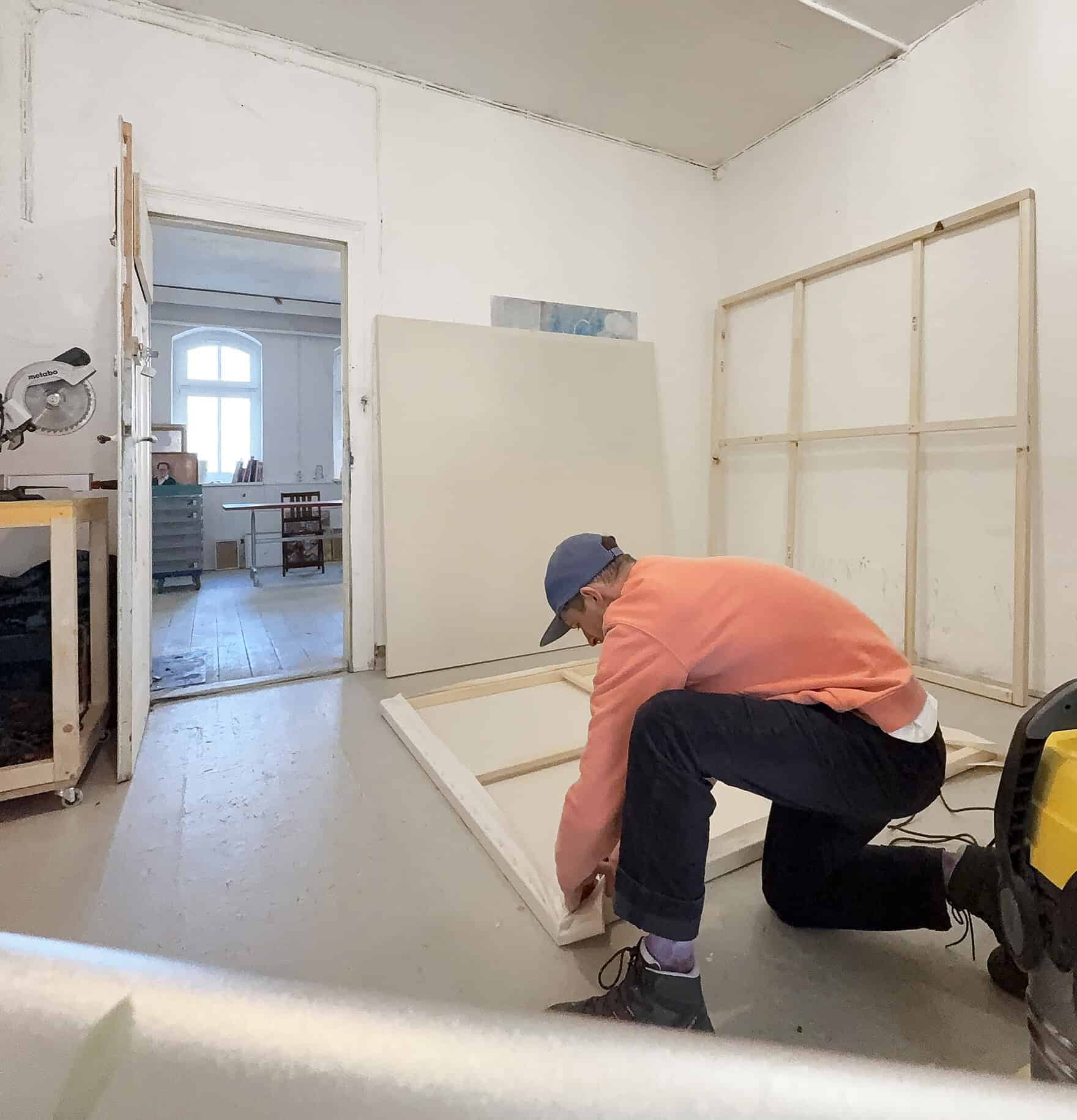 Künstler beim Aufbau eines Rahmens in einem Atelier, Vorbereitung für ein Gemälde, in einem hellen Raum mit weißen Wänden und großen Fenstern.