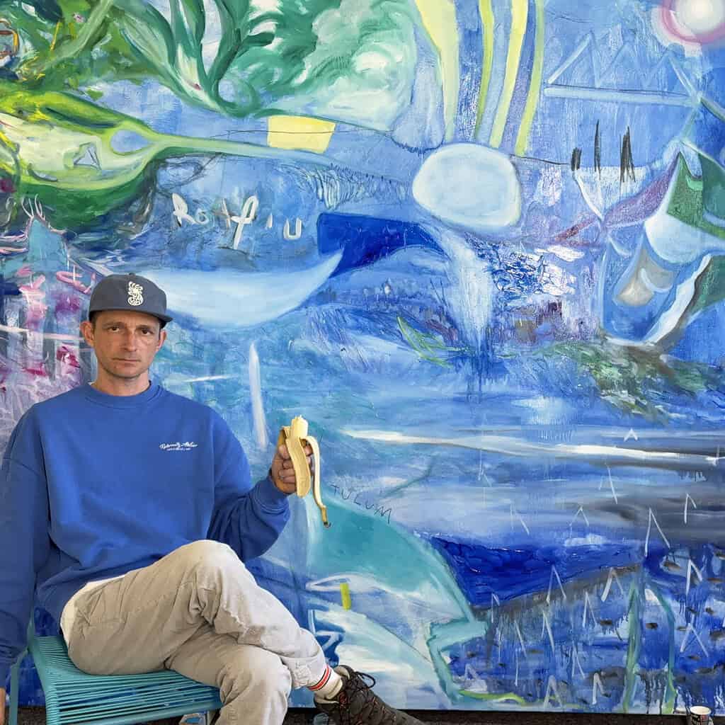 Künstler bei Arbeit vor farbenfrohem Gemälde, das abstrakte und expressive Elemente zeigt.