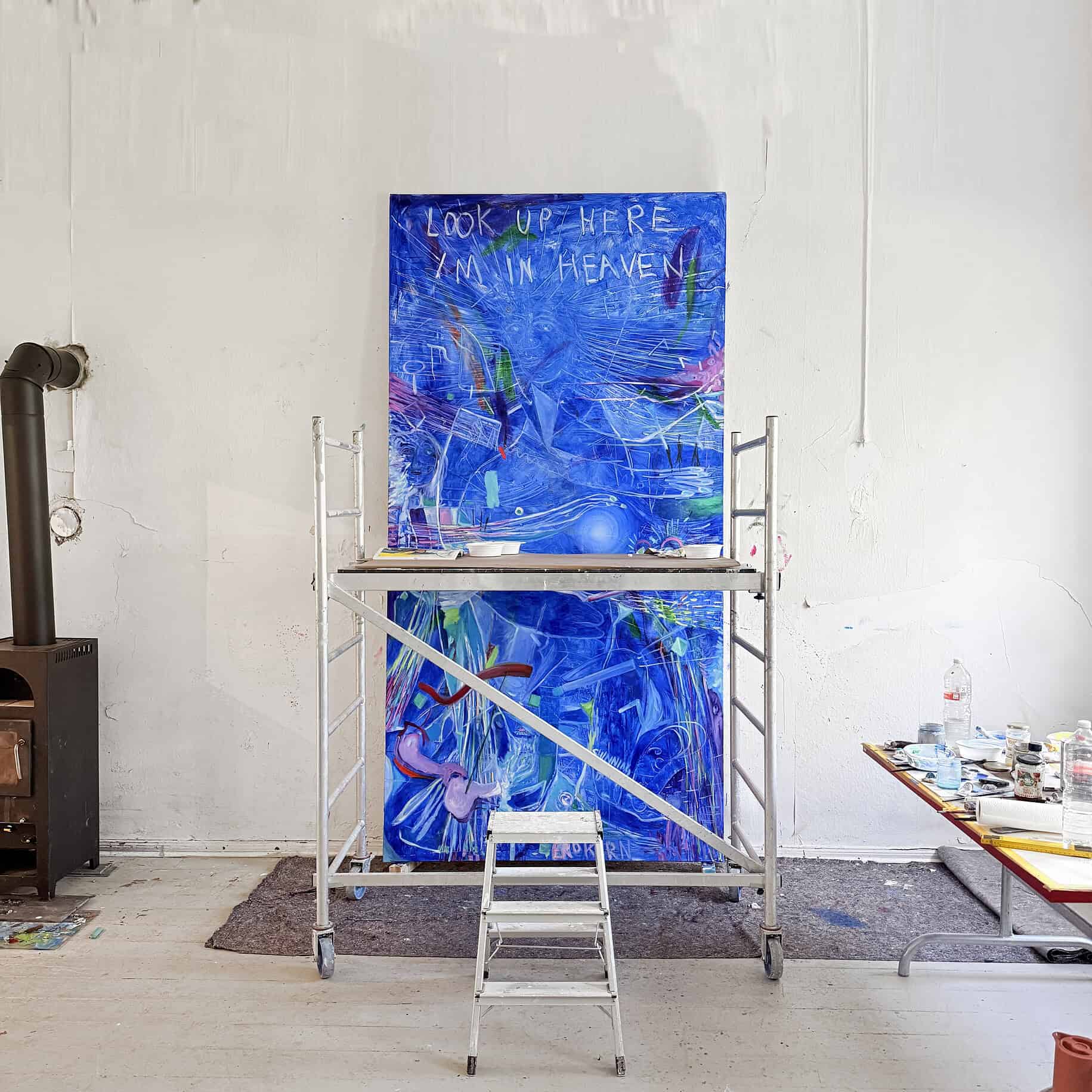 An abstract painting with vibrant colors and the text "Look up here, I'm in heaven" on an easel in an art studio.